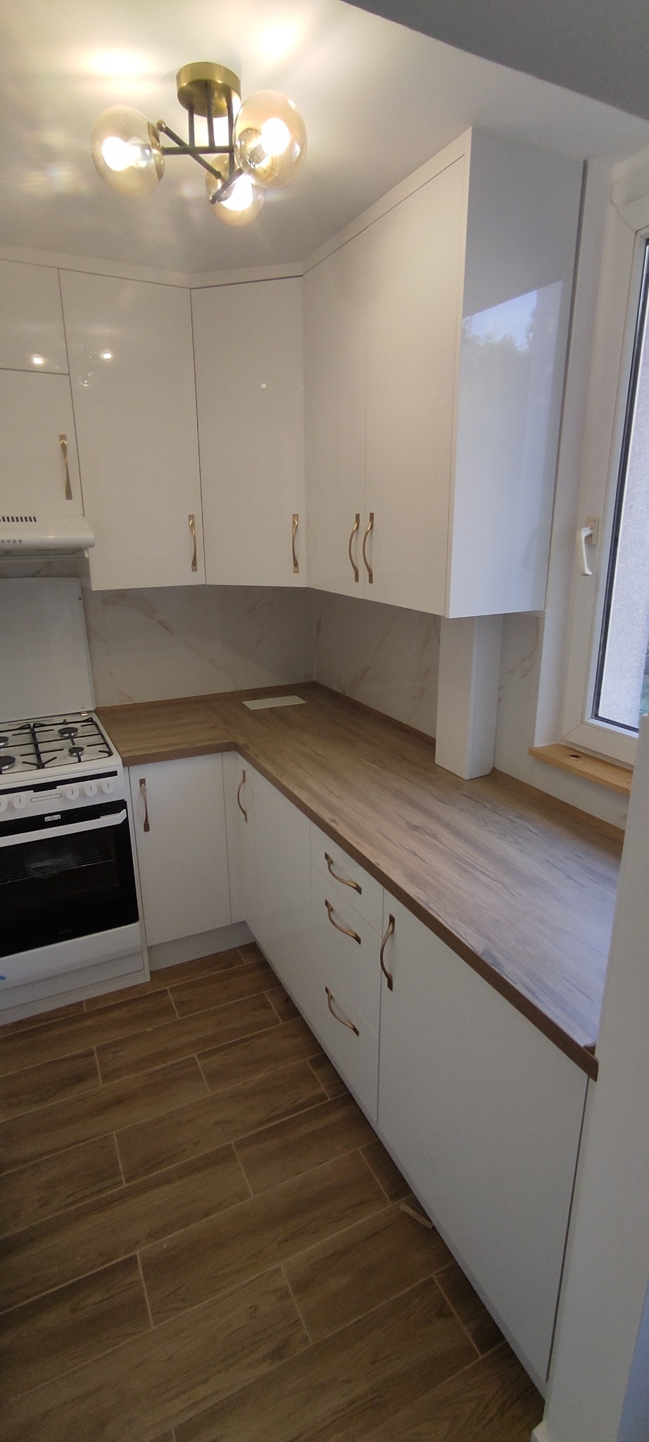 Biała, błyszcząca zabudowa kuchenna z drewnianym blatem, złotymi uchwytami i lampą sufitową z kulistymi kloszami. Widok na kuchenkę i okno.