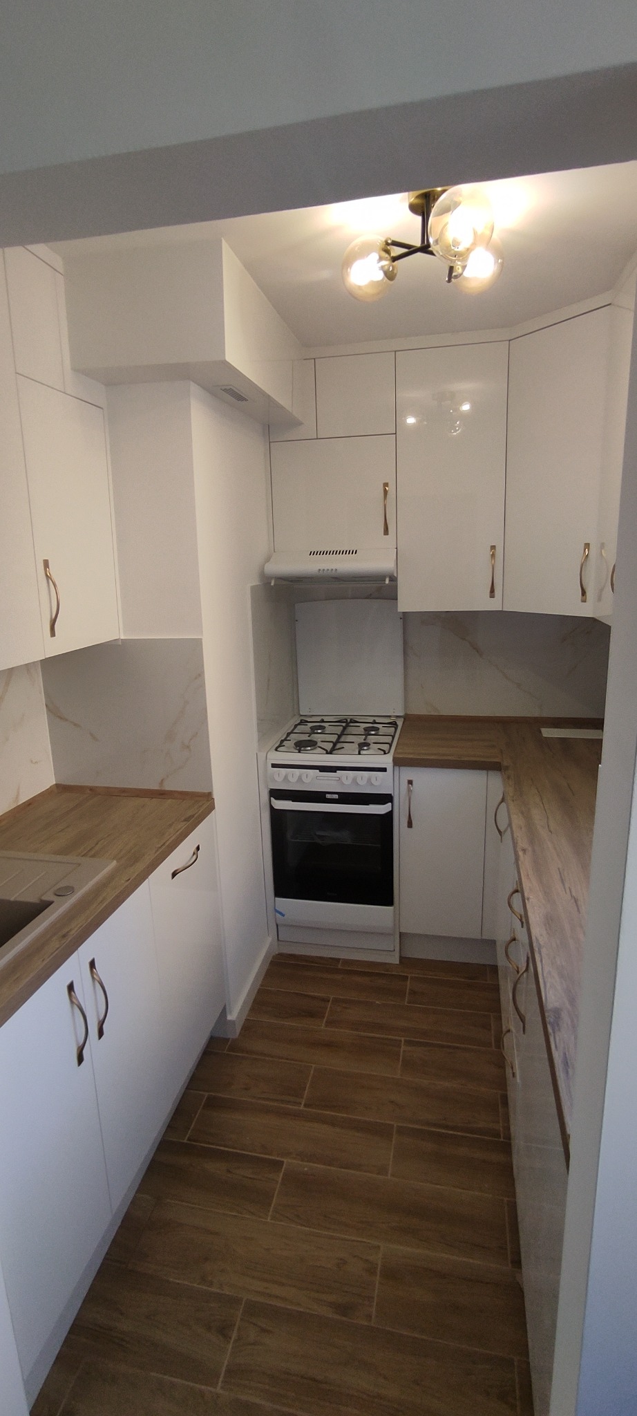 Jasna kuchnia z białymi szafkami i drewnianymi blatami. Widoczna kuchenka gazowa, okap i lampa sufitowa. Uwagę przykuwają złote uchwyty szafek.
