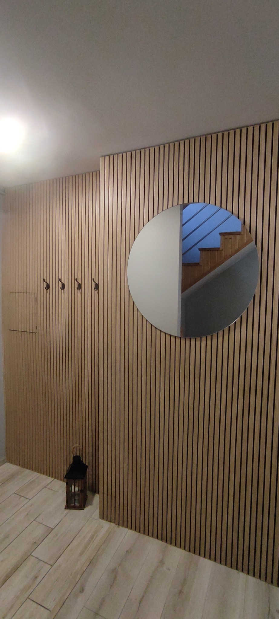 Drewniana ściana z pionowych paneli z lustrem w kształcie koła, w którym odbijają się schody. Dekoracyjna latarenka i wieszaki na ubrania.
