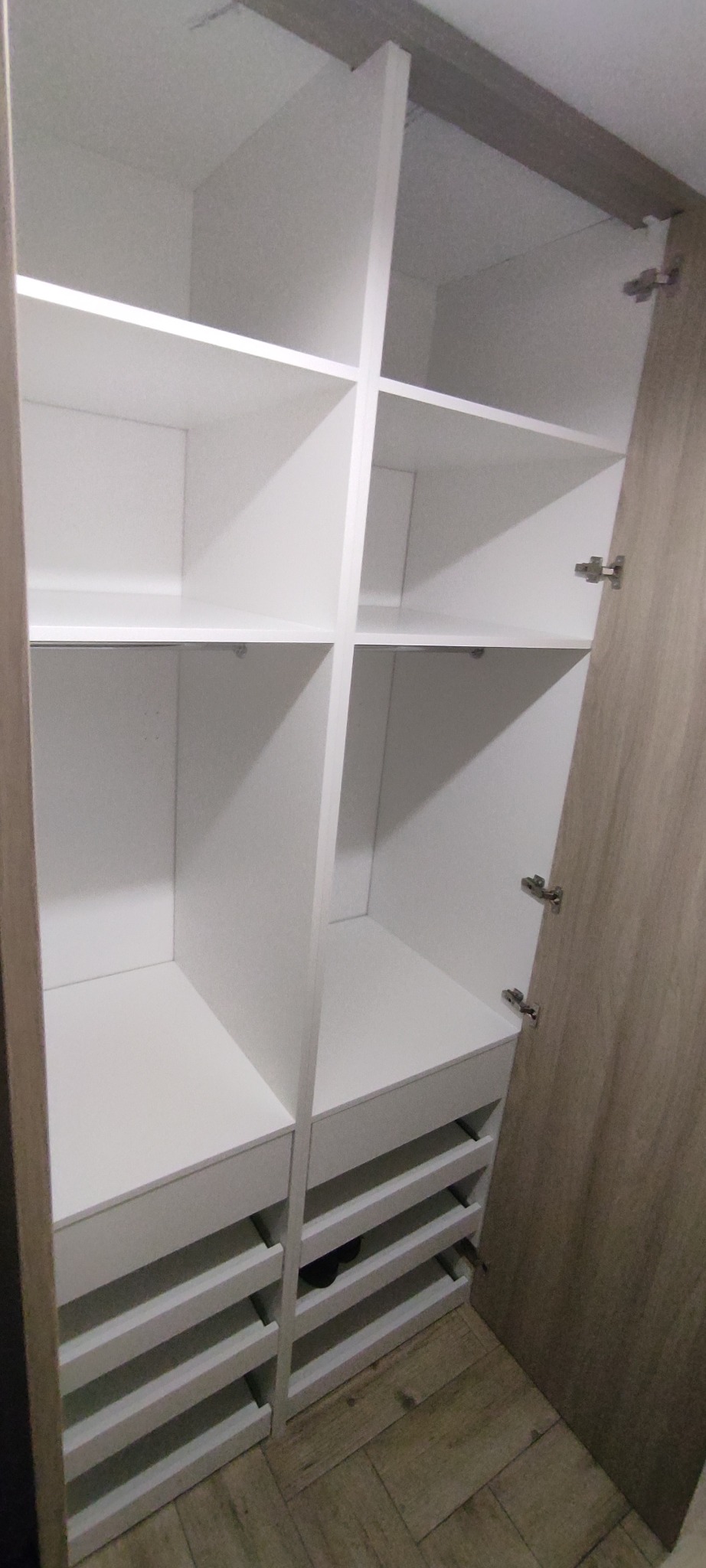 Otwarta, biała szafa wnękowa z półkami i szufladami, widok z bliska. Drzwi szafy w kolorze jasnego drewna. Widoczne zawiasy i podłoga z paneli.