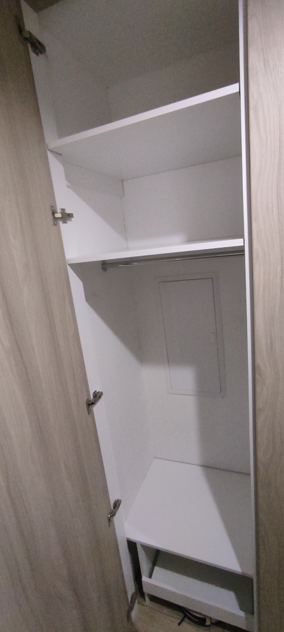 Otwarta szafa wnękowa z półkami i drążkiem na ubrania, widoczne zawiasy na drzwiach, wnętrze białe, boki w kolorze jasnego drewna.