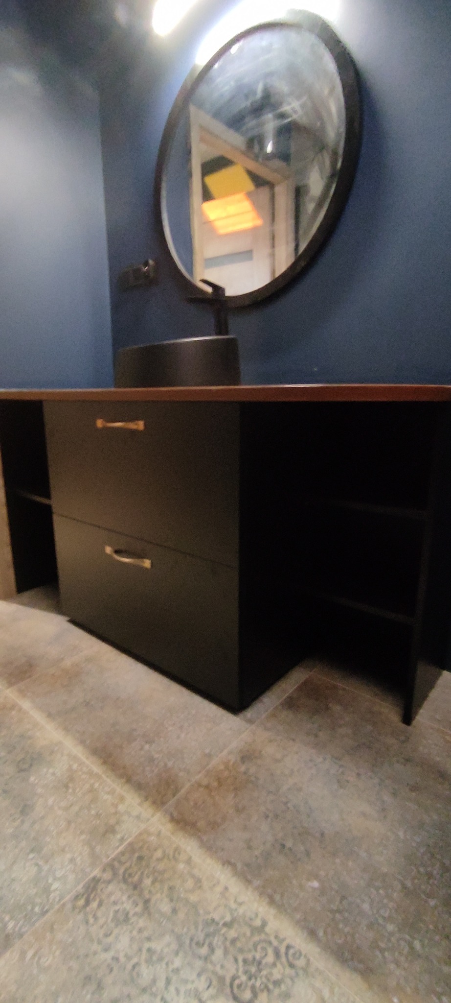 Czarna szafka łazienkowa z umywalką nablatową i okrągłym lustrem na tle ciemnej ściany. Widoczne złote uchwyty i wzorzysta podłoga.