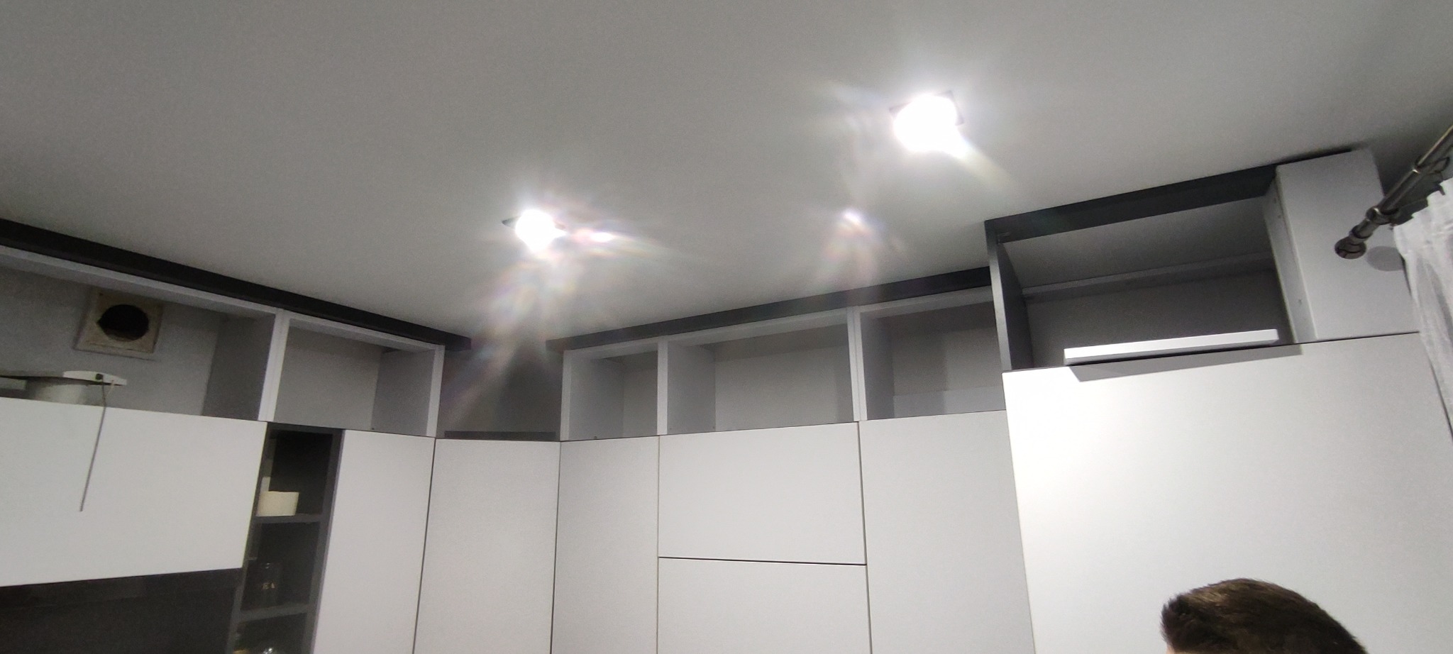 Biała zabudowa kuchenna z czarnymi akcentami, widok od dołu, oświetlenie sufitowe. Górne szafki otwarte, dolne z frontami. Fragment zasłony po prawej.