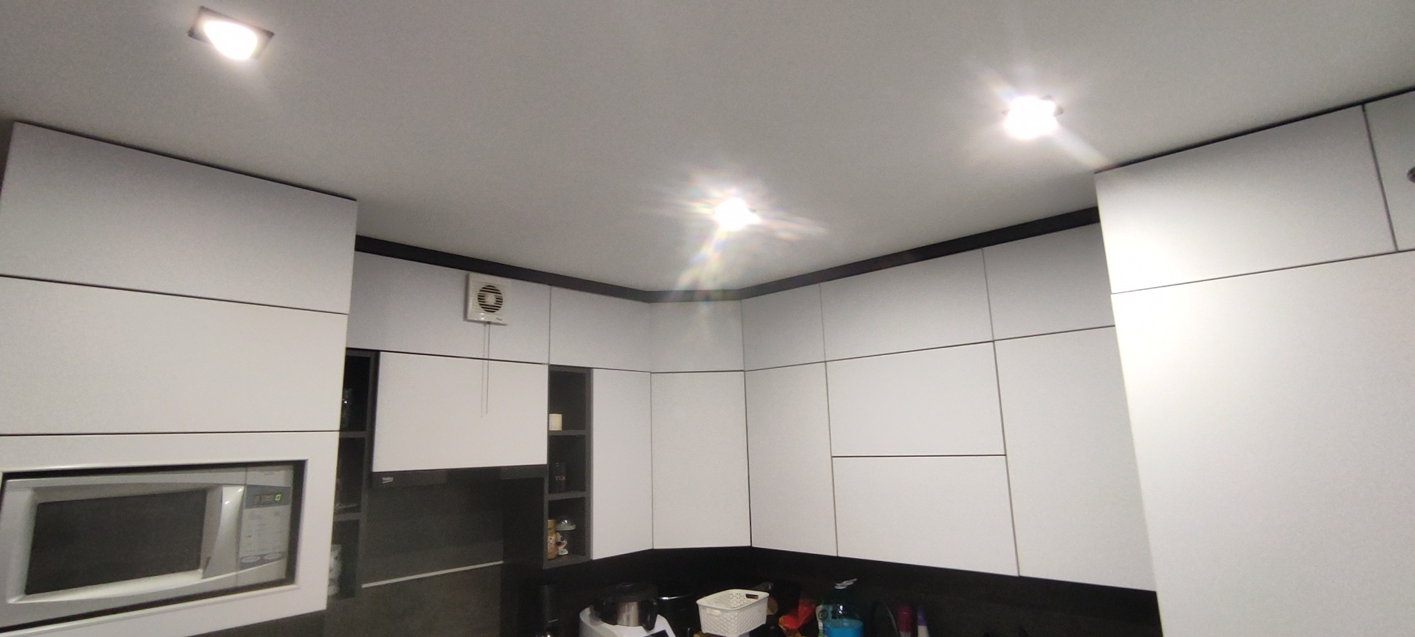 Nowoczesna, biała kuchnia z zabudową pod sufit i czarnymi akcentami. Widoczny fragment blatu i sprzęt AGD. Minimalistyczny design z oświetleniem punktowym.