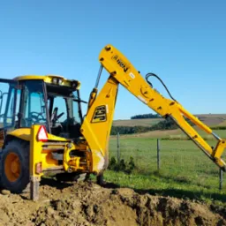 Żółta koparko-ładowarka JCB w trakcie prac ziemnych na tle zielonej łąki i błękitnego nieba. Widoczny wykop i otwarte drzwi kabiny.