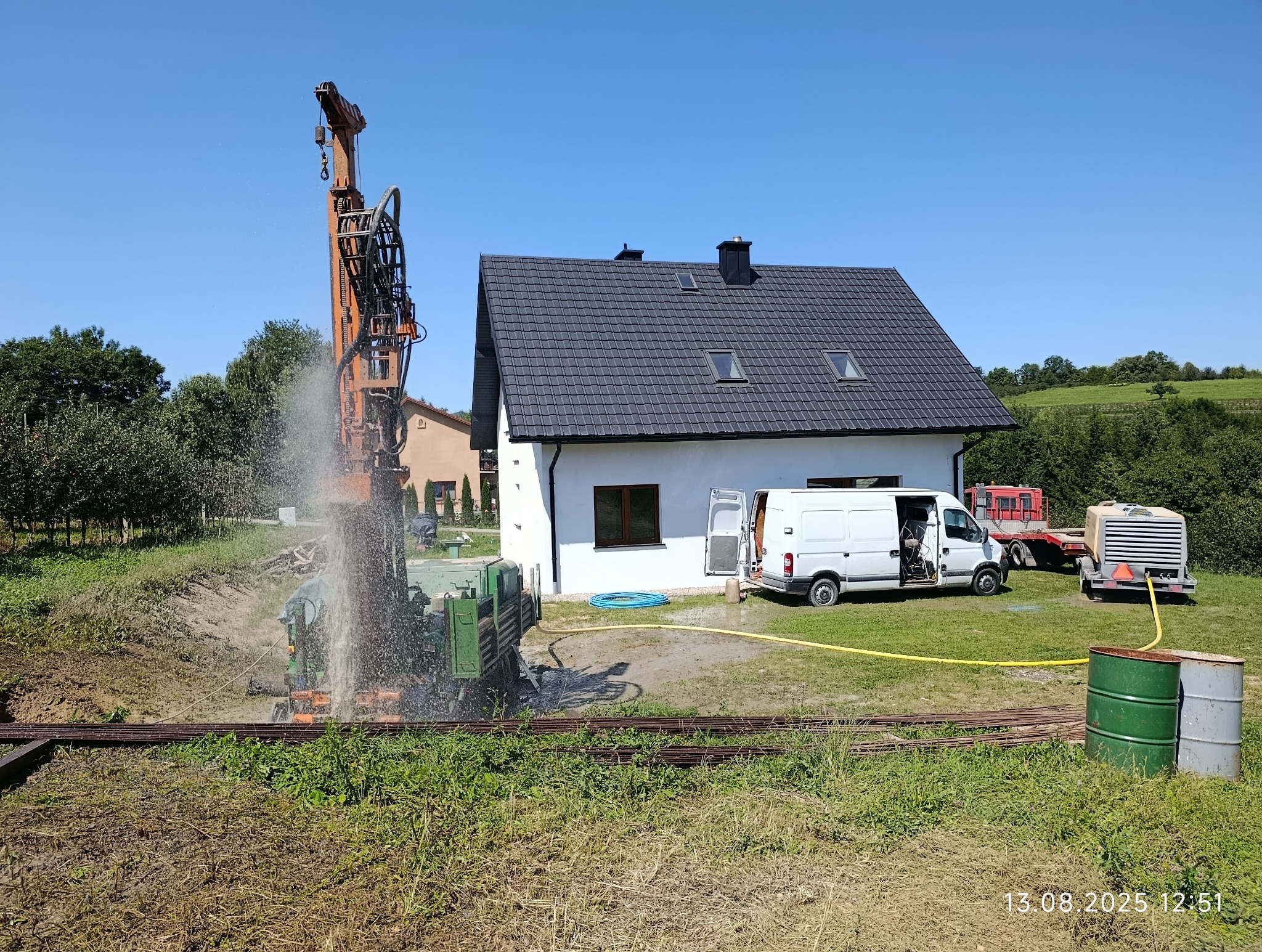 Wiercenie studni głębinowej w Florynka. Wiertnica w akcji, woda tryska spod ziemi. Biały dom w tle, sprzęt i narzędzia na trawie. Błękitne niebo.