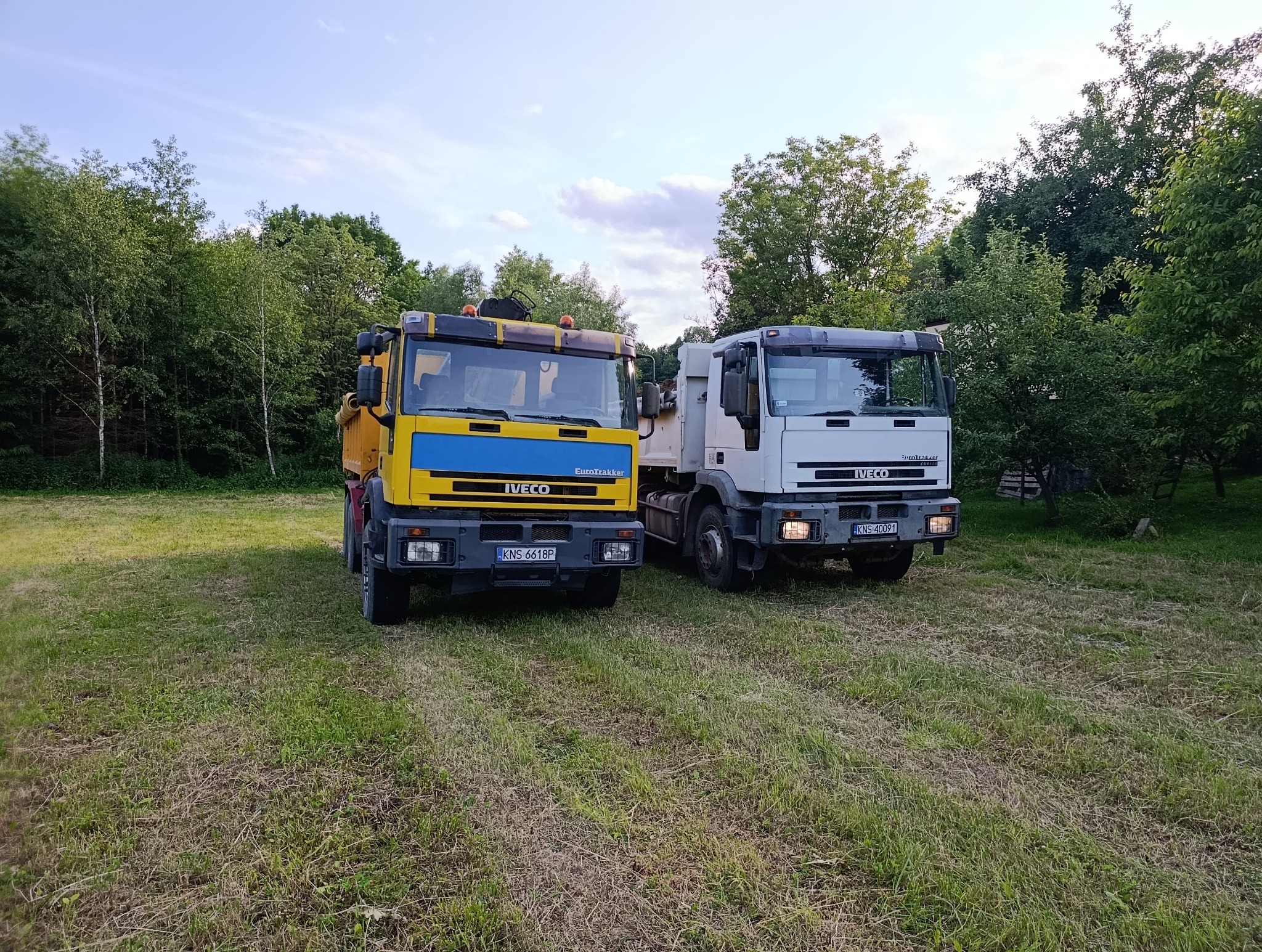 Dwa samochody ciężarowe marki Iveco, żółto-niebieski i biały, zaparkowane na trawiastym terenie. Widoczne tablice rejestracyjne i fragmenty lasu w tle.