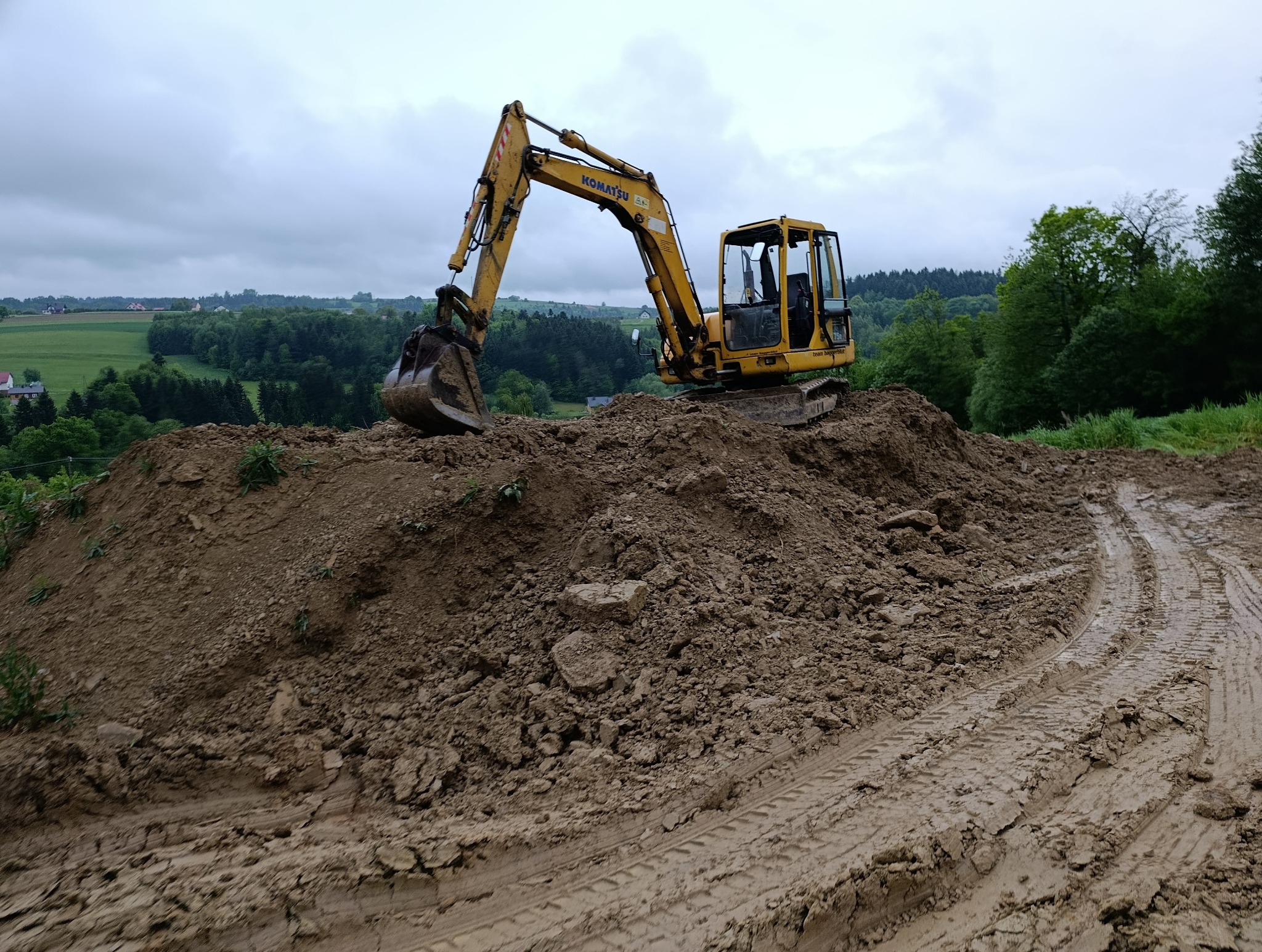 Żółta koparka Komatsu na stercie ziemi z widocznymi śladami opon, w tle zielony krajobraz i pochmurne niebo. Prace ziemne w toku.