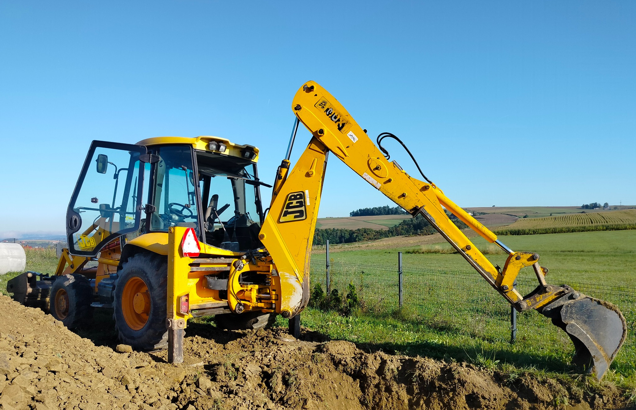 Żółta koparko-ładowarka JCB w trakcie prac ziemnych na tle zielonej łąki i błękitnego nieba. Widoczny wykop i otwarte drzwi kabiny.
