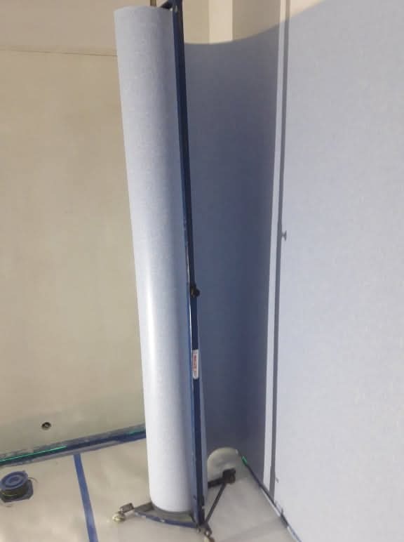 Rolka jasnej wykładziny w specjalnym stojaku na kółkach, przygotowana do montażu w pomieszczeniu z zabezpieczonymi taśmą krawędziami podłogi.