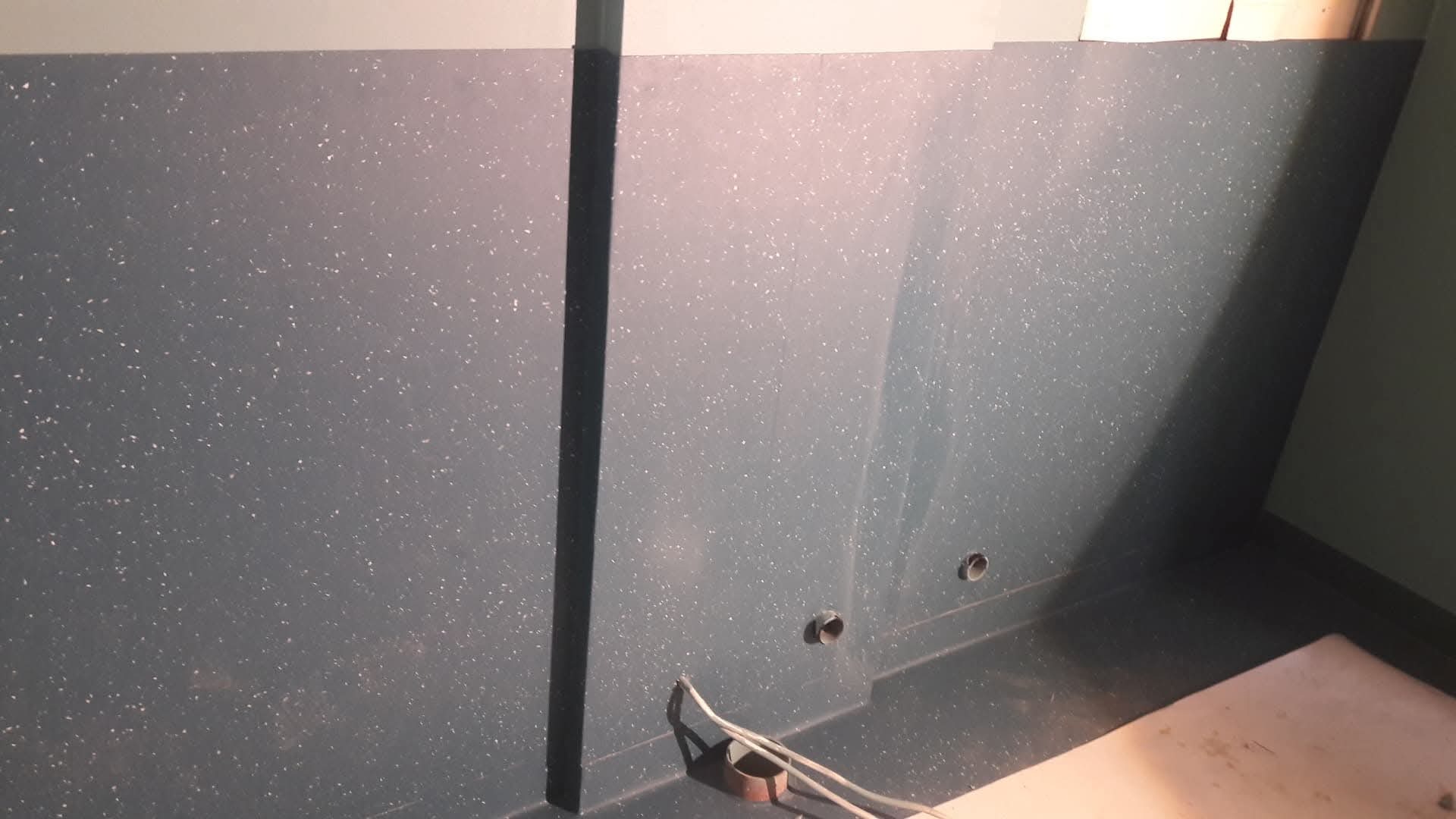 Szara wykładzina PCV z białymi drobnymi plamkami na ścianie i podłodze, widoczne dwa otwory w ścianie, listwa przypodłogowa i fragment przewodów na podłodze.