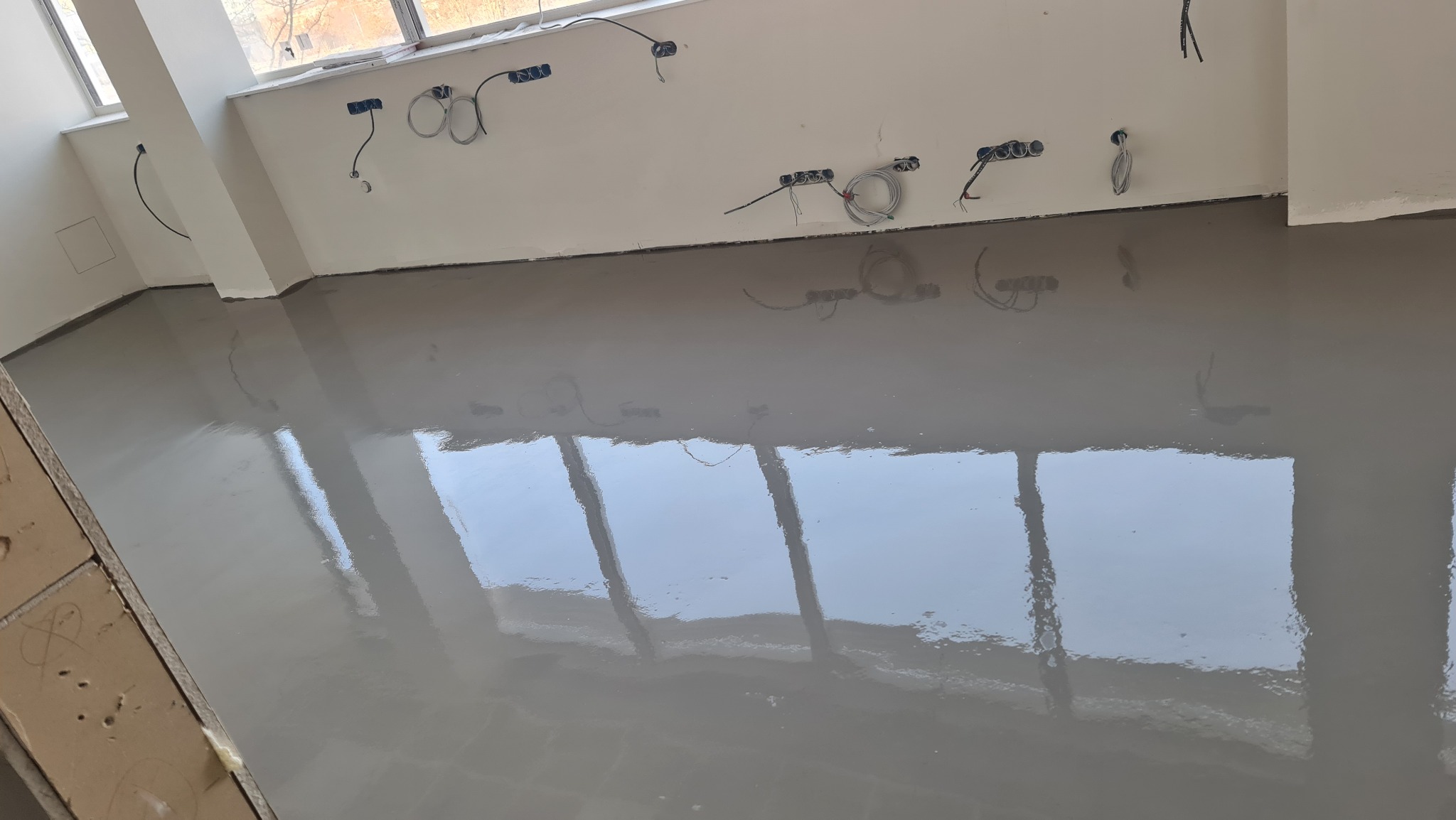 Świeżo wylana, samopoziomująca wylewka podłogowa w jasnym, pustym pomieszczeniu z widocznymi przewodami elektrycznymi na ścianie.