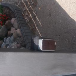 Bramy Kali Bogumił Kalisciak - Szary słupek bramy z zamontowanym automatem NICE, obok rabata z kamieniami i kwiatami, cień osoby w prawym górnym rogu.