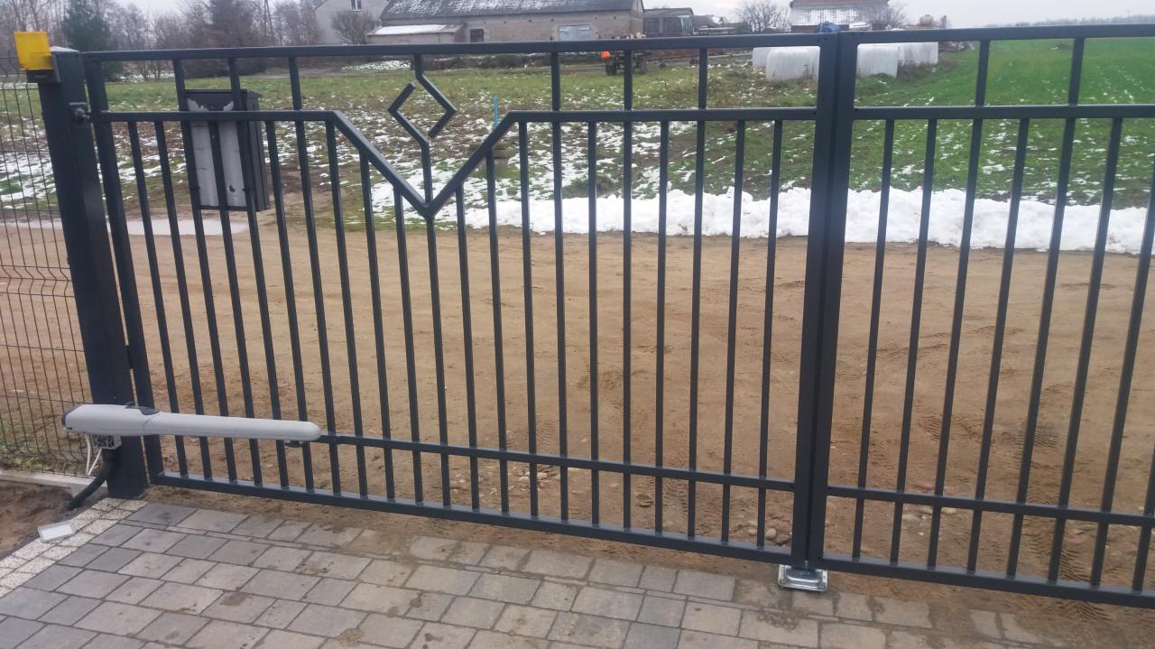 Nowoczesna, metalowa brama wjazdowa z automatycznym napędem, widoczna na tle zaśnieżonego krajobrazu i budynków gospodarczych.