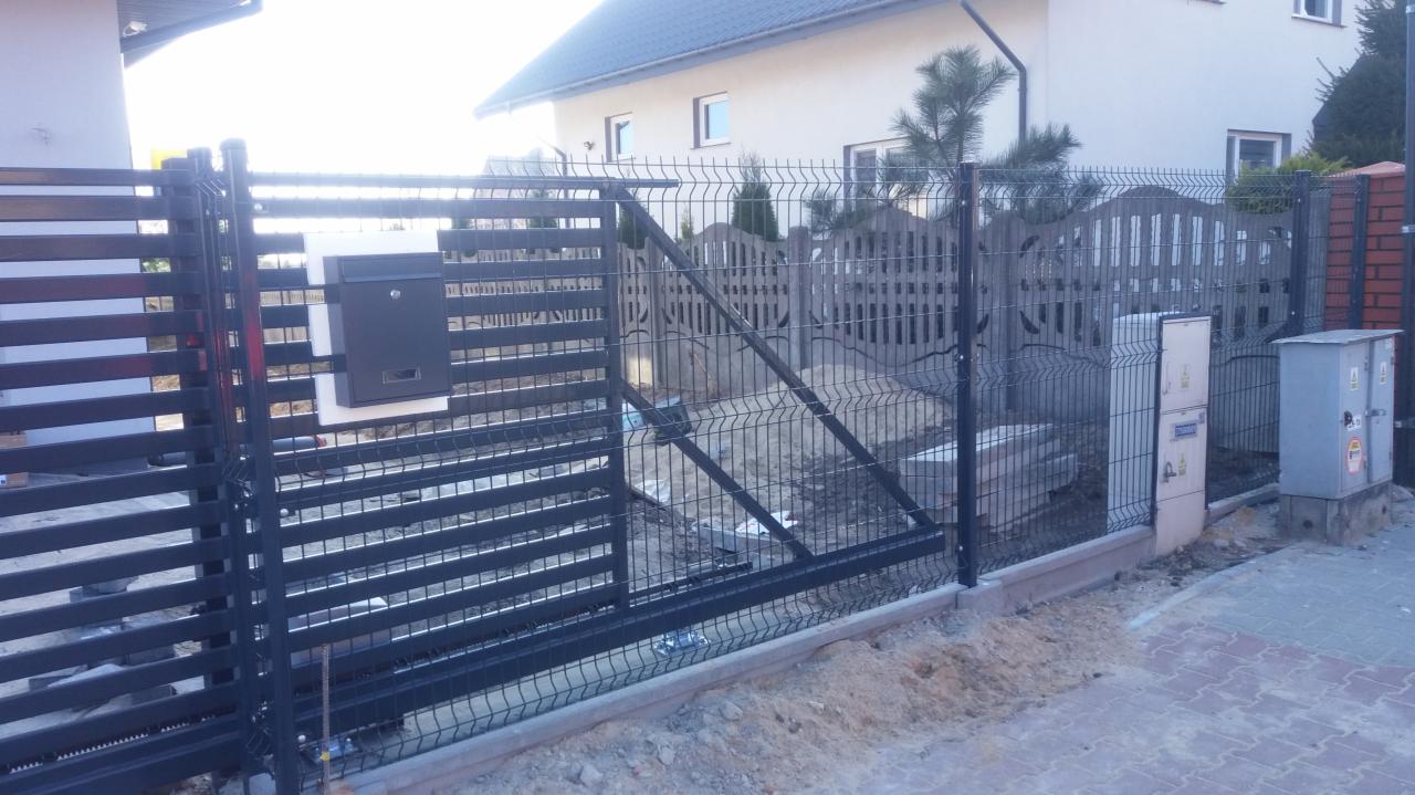 Nowoczesne ogrodzenie panelowe z przesuwną bramą i skrzynką na listy, widoczne także elementy automatyki bramowej i fragment ogrodzenia betonowego w tle.