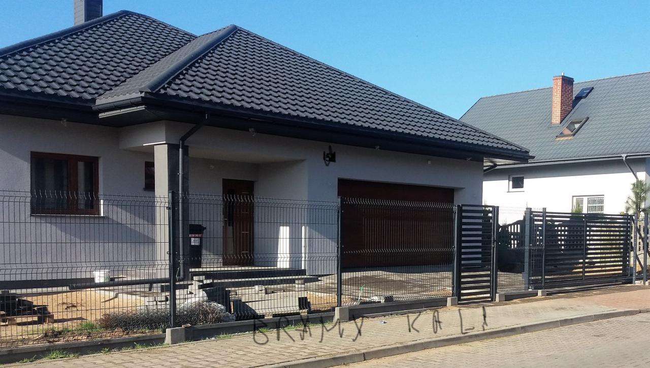 Nowoczesny dom jednorodzinny z szarą elewacją, grafitową dachówką i czarnym ogrodzeniem panelowym oraz bramą przesuwną, widoczne przygotowania do dalszych prac montażowych.
