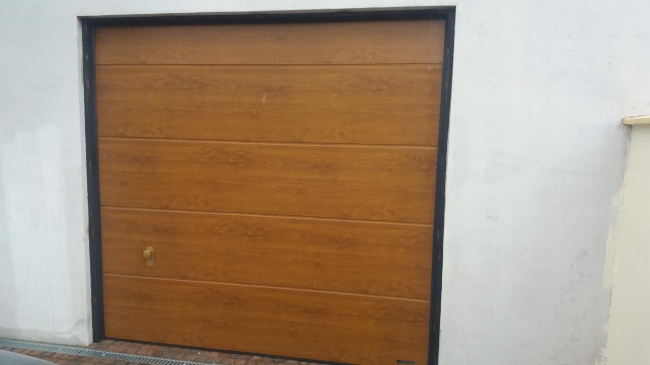 Segmentowa brama garażowa w kolorze drewna, z czarną ramą i prostym uchwytem, zamontowana w białym, surowym murze.