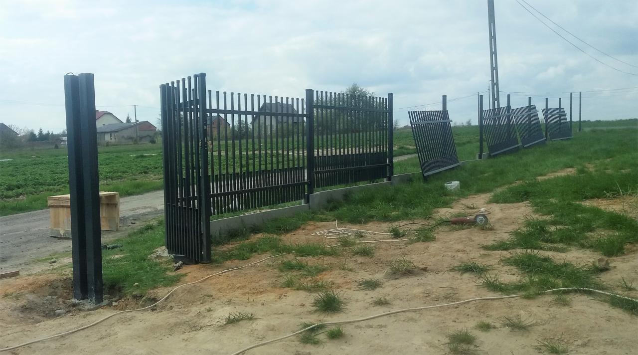 Częściowo zamontowane czarne ogrodzenie metalowe z pionowymi prętami, widoczne słupki, podmurówka, rozłożone panele, szlifierka kątowa i przewód zasilający na trawiastym terenie.