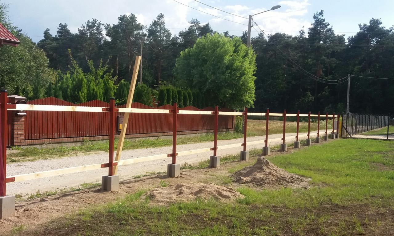 Wznoszenie ogrodzenia panelowego: czerwone słupki z betonowymi podstawami i drewnianymi poprzeczkami na tle zieleni i częściowo widocznego domu.