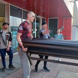 Comfort LOGISTYK twój dom Krzysztof Funk - Trzej mężczyźni przenoszą ciemnobrązową konstrukcję metalową, prawdopodobnie ławkę lub stelaż, przed budynkiem z czerwonymi i szarymi elementami, widoczna kobieta obserwująca.