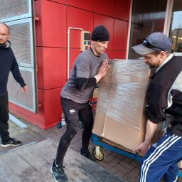 Comfort LOGISTYK twój dom Krzysztof Funk - Trzech mężczyzn transportuje duży, owinięty folią karton na wózku transportowym przed budynkiem o czerwonej elewacji.