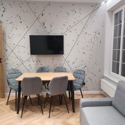 Comfort LOGISTYK twój dom Krzysztof Funk - Jasne wnętrze jadalni z drewnianym stołem, szarymi krzesłami z pikowaniem, telewizorem na ścianie z dekoracyjnym wzorem i parkietem w jodełkę.