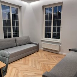 Comfort LOGISTYK twój dom Krzysztof Funk - Jasne wnętrze pokoju z parkietem w jodełkę, dwoma szarymi sofami, drewnianym stołem z szarymi krzesłami i dwoma oknami z widokiem na budynki nocą.