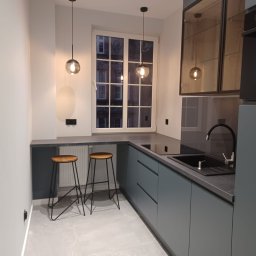 Comfort LOGISTYK twój dom Krzysztof Funk - Nowoczesna kuchnia w odcieniach szarości z dwoma hokerami przy blacie, oświetlona wiszącymi lampami kulami, z widokiem na kamienicę za oknem.