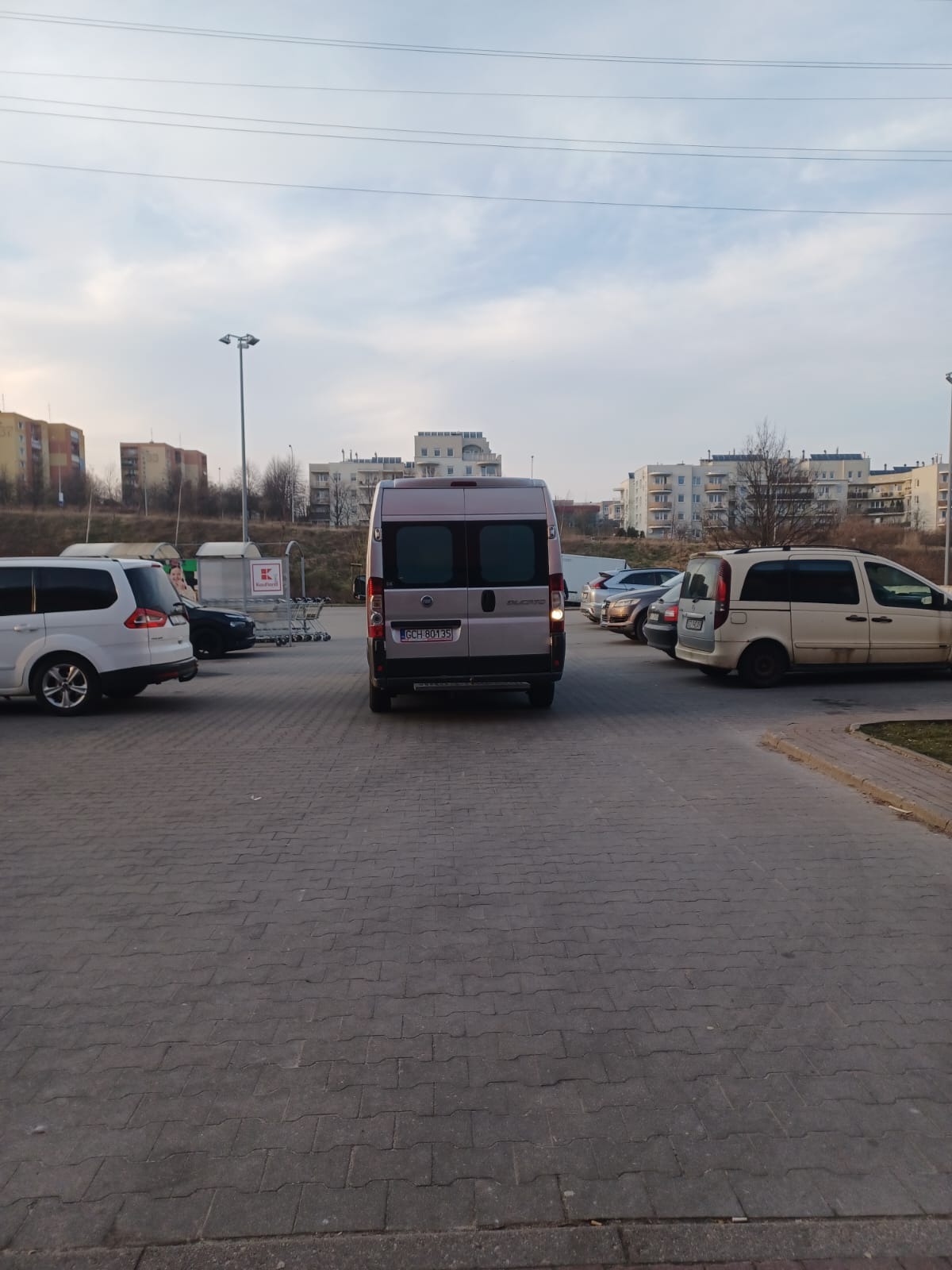 Szary furgon zaparkowany na parkingu sklepu, widziany od tyłu, z włączonymi światłami stopu, sugerujący gotowość do wyjazdu.