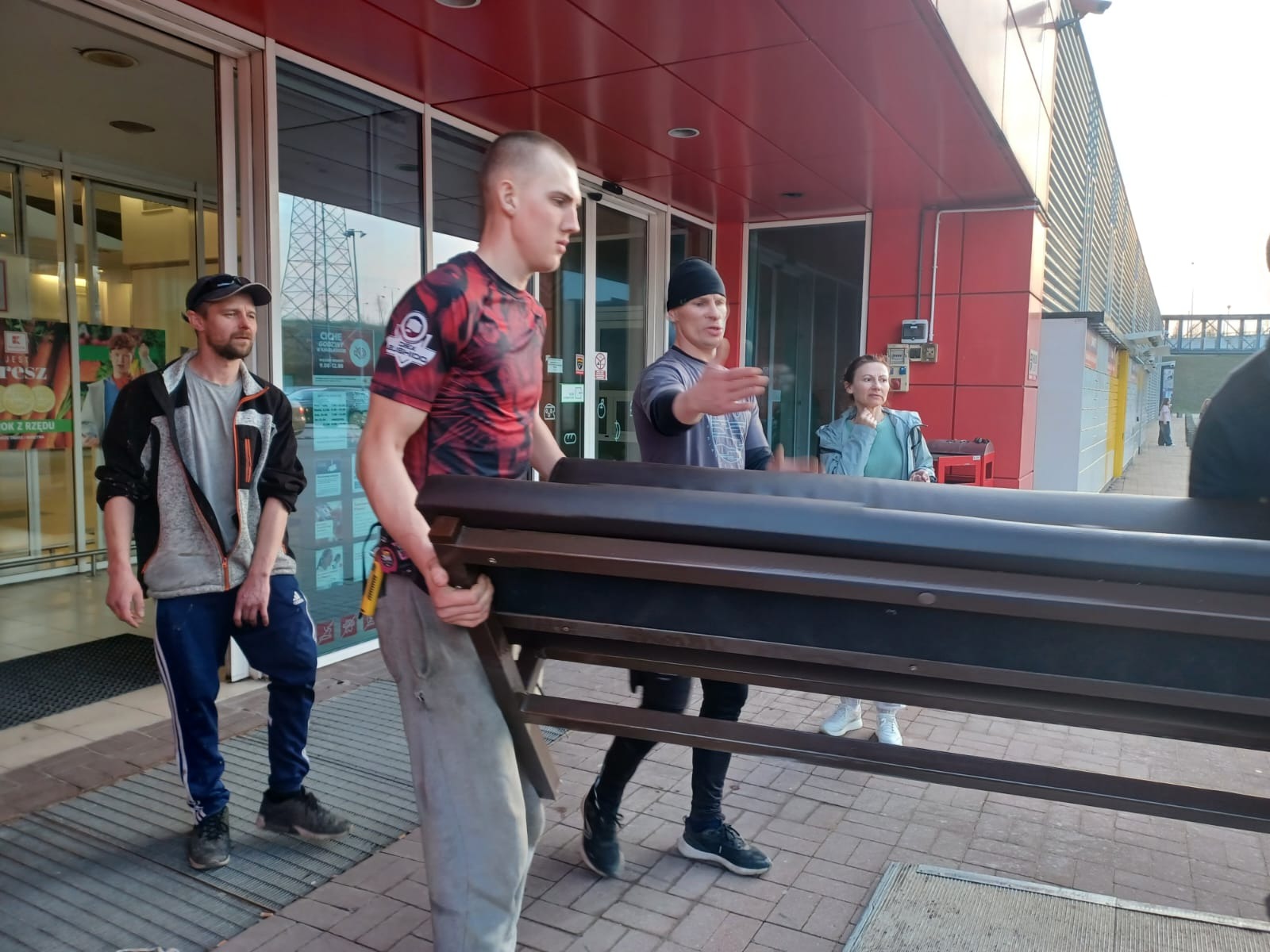 Trzej mężczyźni przenoszą ciemnobrązową konstrukcję metalową, prawdopodobnie ławkę lub stelaż, przed budynkiem z czerwonymi i szarymi elementami, widoczna kobieta obserwująca.