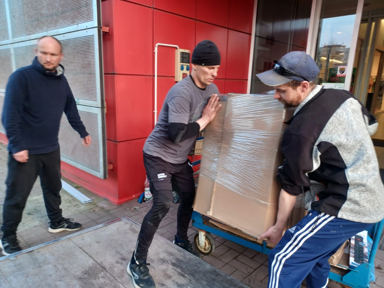 Trzech mężczyzn transportuje duży, owinięty folią karton na wózku transportowym przed budynkiem o czerwonej elewacji.
