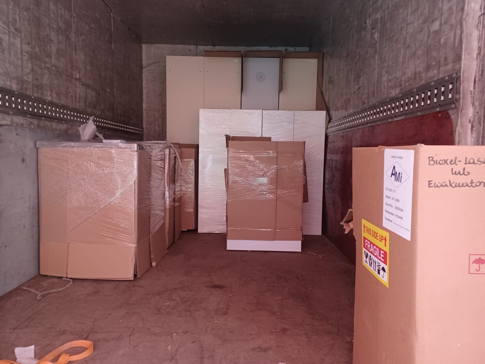 Wnętrze skrzyni ładunkowej wypełnionej meblami, niektóre owinięte folią stretch, inne w kartonach, gotowe do transportu. Na jednym z kartonów widoczna naklejka ostrzegawcza 'Fragile' oraz opis...