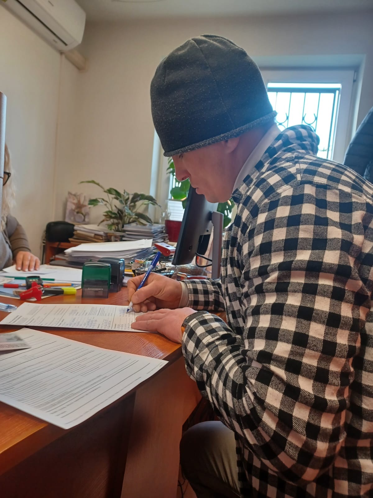 Mężczyzna w czarno-białej koszuli w kratę i szarej czapce podpisuje dokument na biurku w biurze, widoczne pieczątki i inne dokumenty.