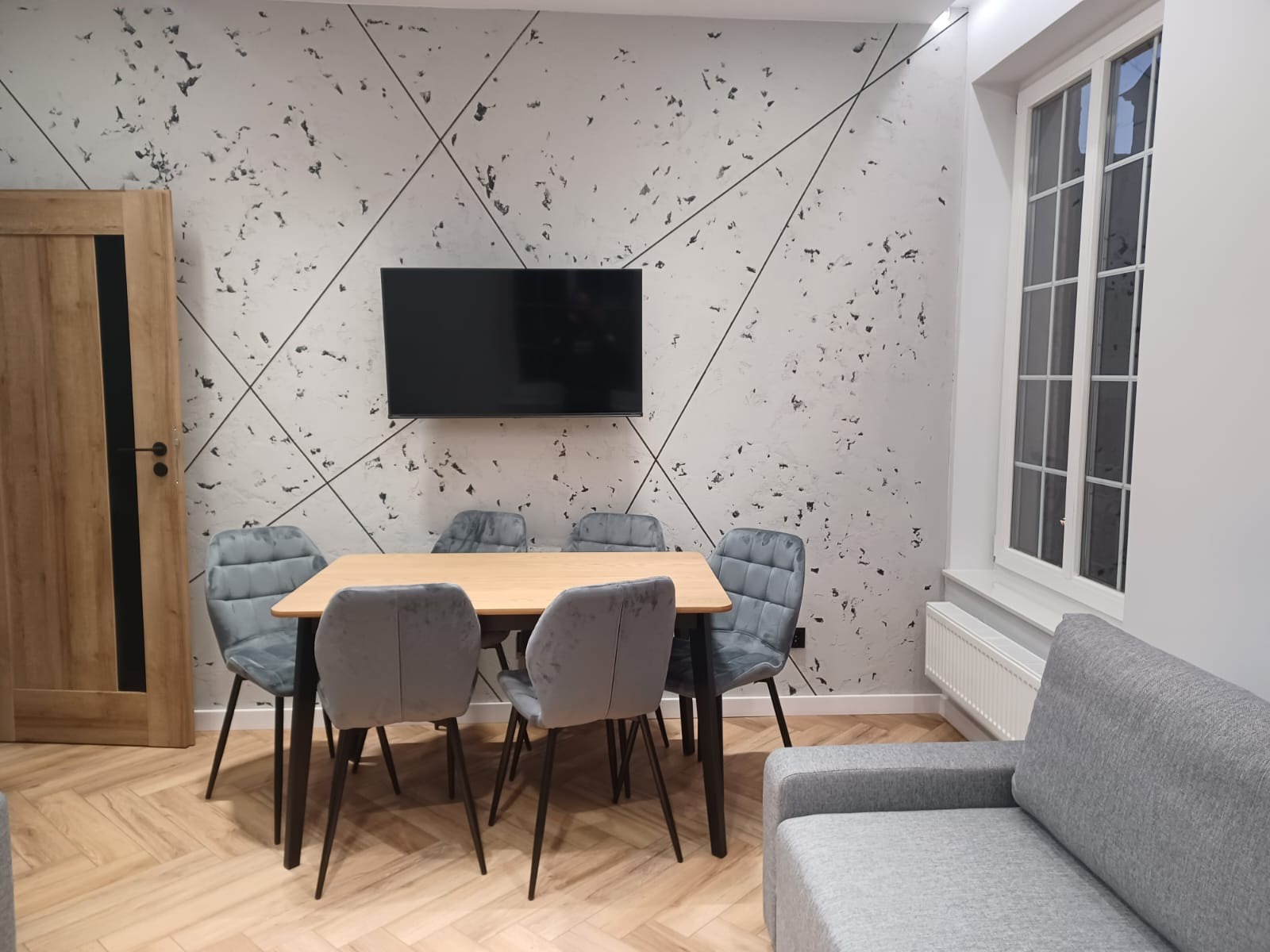 Jasne wnętrze jadalni z drewnianym stołem, szarymi krzesłami z pikowaniem, telewizorem na ścianie z dekoracyjnym wzorem i parkietem w jodełkę.