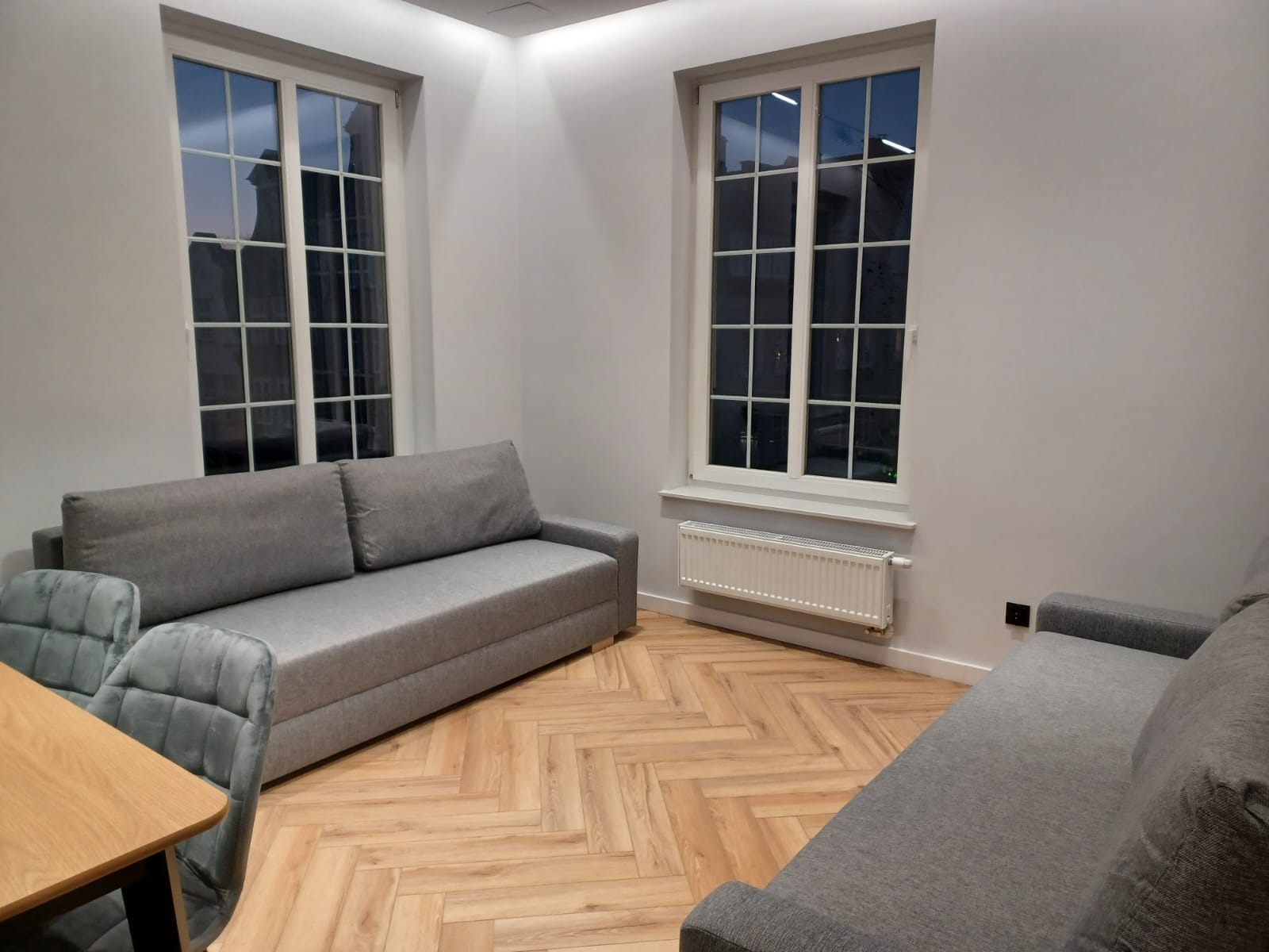 Jasne wnętrze pokoju z parkietem w jodełkę, dwoma szarymi sofami, drewnianym stołem z szarymi krzesłami i dwoma oknami z widokiem na budynki nocą.