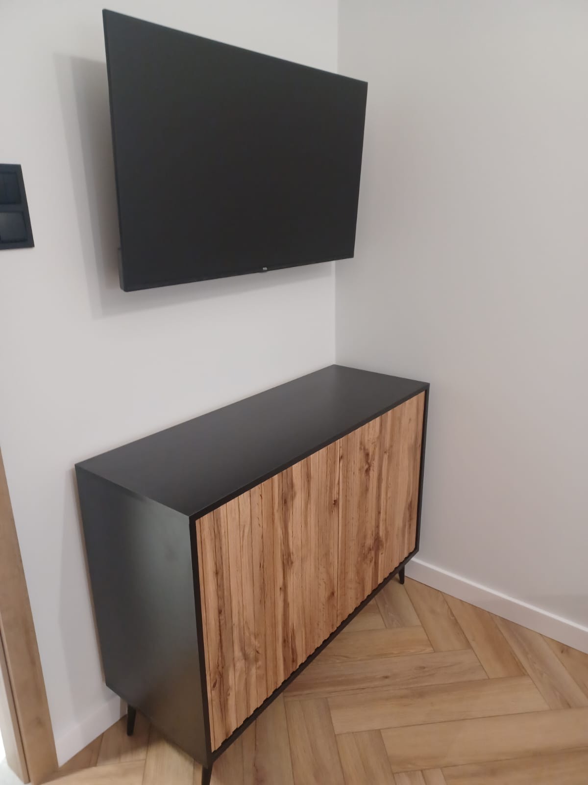Nowoczesna szafka RTV z drewnianymi lamelami i czarnym blatem, zawieszony telewizor na białej ścianie, parkiet ułożony w jodełkę.