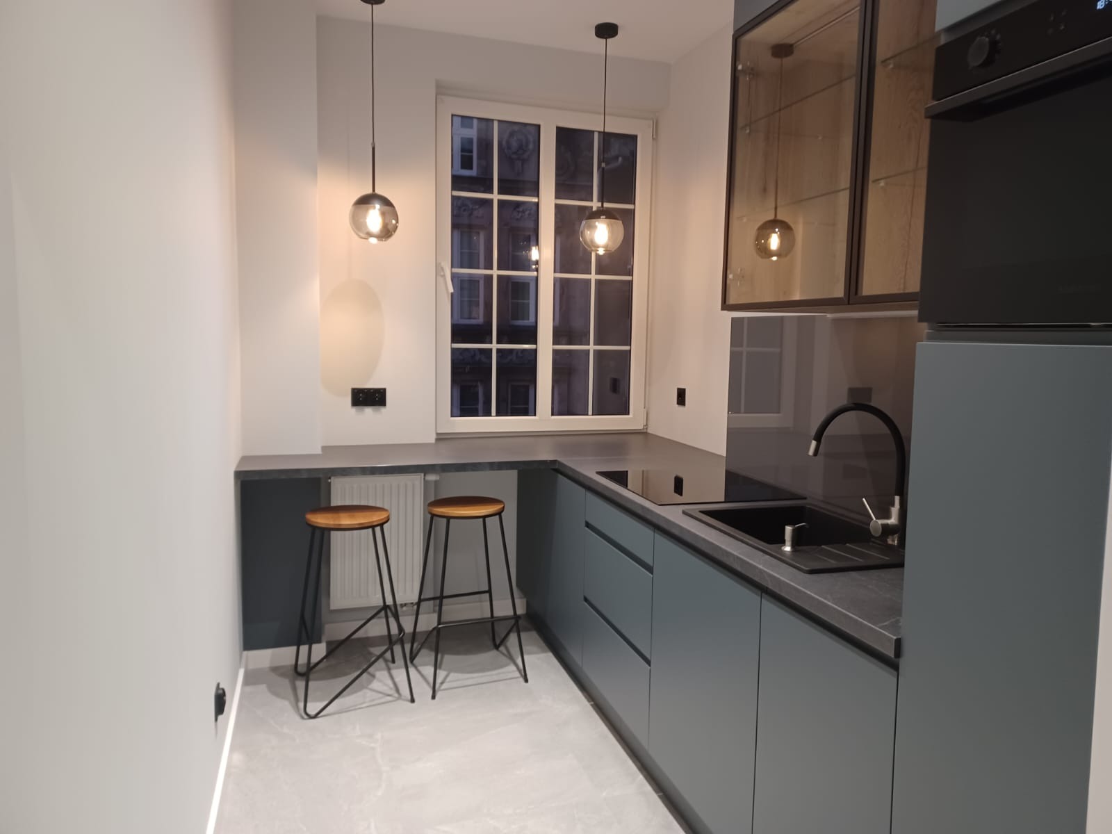 Nowoczesna kuchnia w odcieniach szarości z dwoma hokerami przy blacie, oświetlona wiszącymi lampami kulami, z widokiem na kamienicę za oknem.