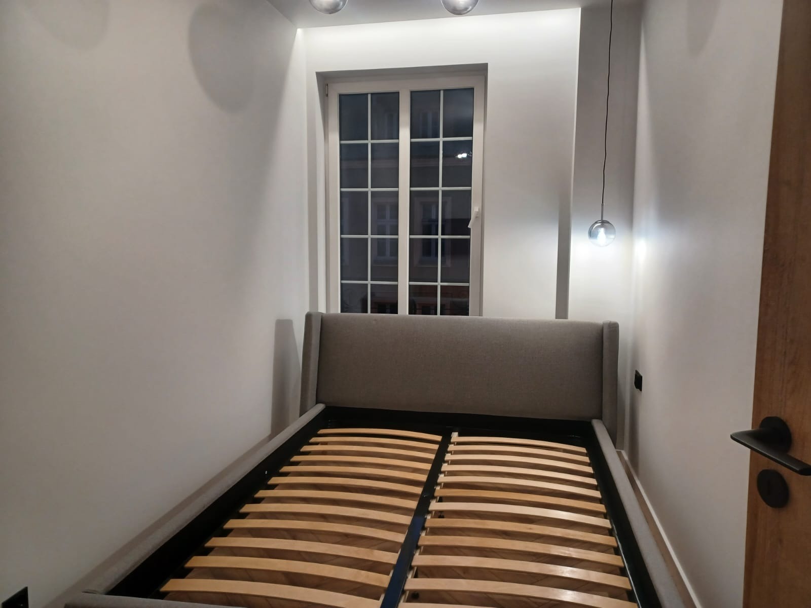 Stelaż łóżka z drewnianymi listwami w szarej ramie z zagłówkiem, widok z boku na tle białej ściany z oknem i wiszącą lampą.