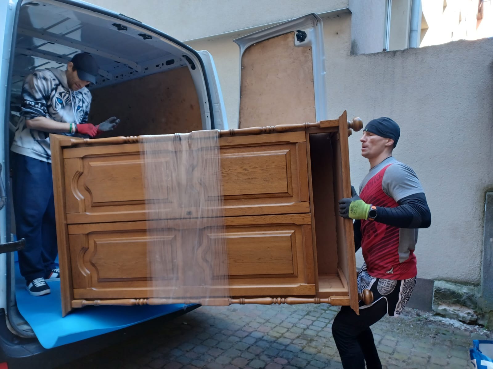 Dwóch mężczyzn przenosi owiniętą folią komodę z szufladami do białego busa. Jeden z mężczyzn ma na sobie czapkę z daszkiem i bluzę w tygrysy, drugi czarną czapkę i sportową koszulkę.