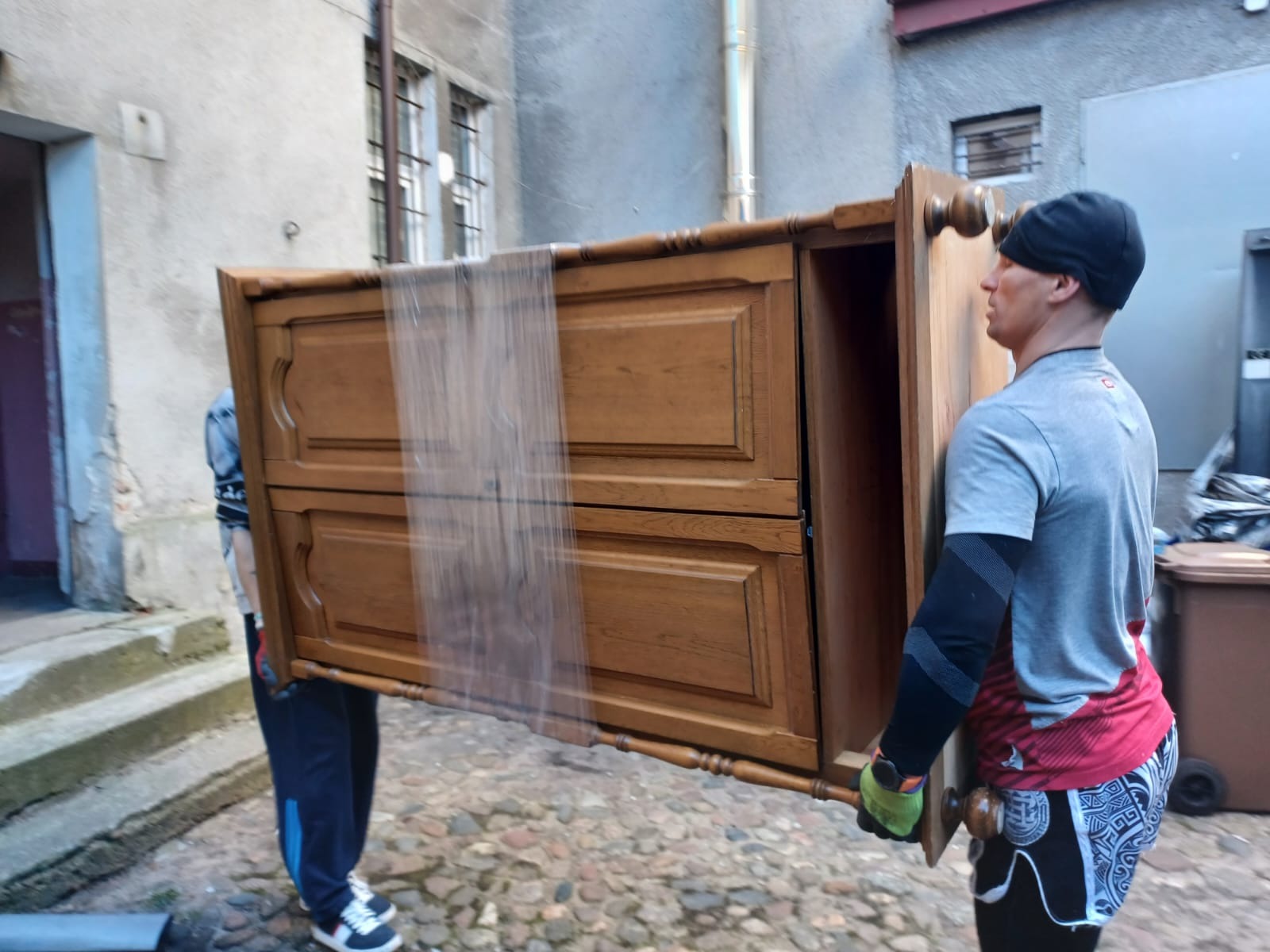 Dwóch mężczyzn przenosi starą, drewnianą komodę zabezpieczoną folią, na tle dziedzińca kamienicy z brukowaną nawierzchnią.