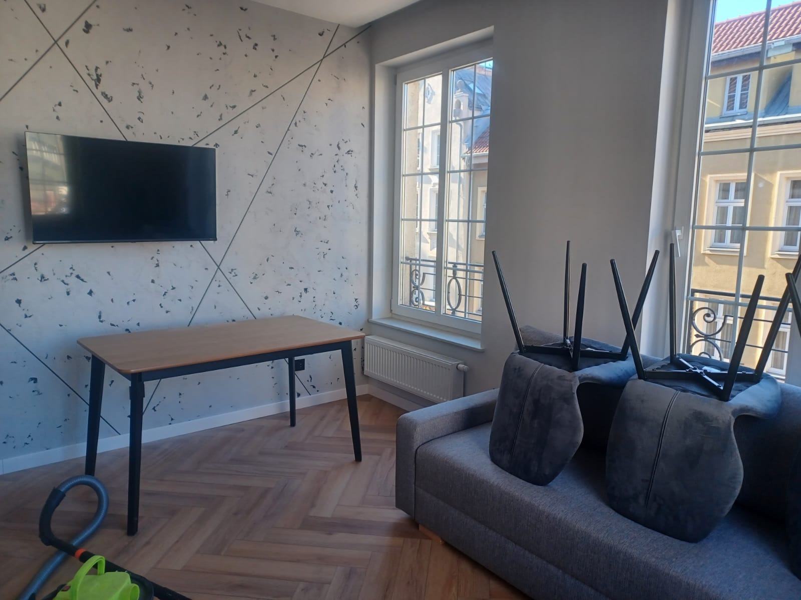 Salon w Luzinie przed sprzątaniem: odkurzacz na podłodze, stół i odwrócone krzesła na szarej kanapie, ściana z geometrycznym wzorem.