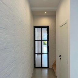 Geso Steel - Wnętrze korytarza z czarną, przeszkloną ścianką działową, imitującą drzwi, na końcu; ściana po lewej stronie z fakturą imitującą kamień; minimalistyczne oświetlenie sufitowe.