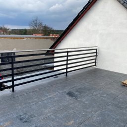 Geso Steel - Czarna, pozioma balustrada na tarasie wykończonym szarymi płytkami, widok na budynki w tle i pochmurne niebo.