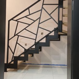 Geso Steel - Nowoczesne schody z drewnianymi stopniami i geometryczną, czarną balustradą, widziane z dołu przez otwarte drzwi. Biała podłoga i jasna ściana w tle.