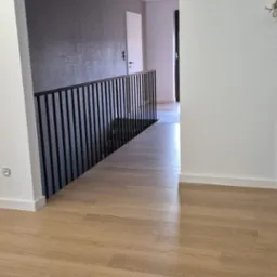 Nowoczesne wnętrze z drewnianą podłogą i czarną balustradą schodową. Szara ściana w tle dodaje elegancji. Widok z góry na korytarz.