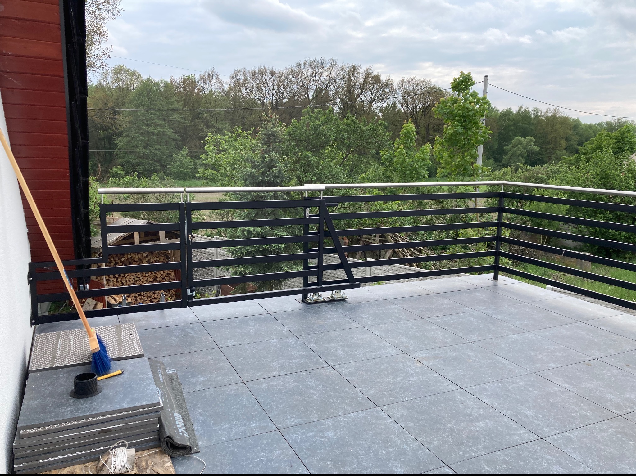 Montaż balustrady balkonowej z czarnych, poziomych profili na tle zieleni, widoczne płytki podłogowe i narzędzia pracy: miotła i nóż do tapet.