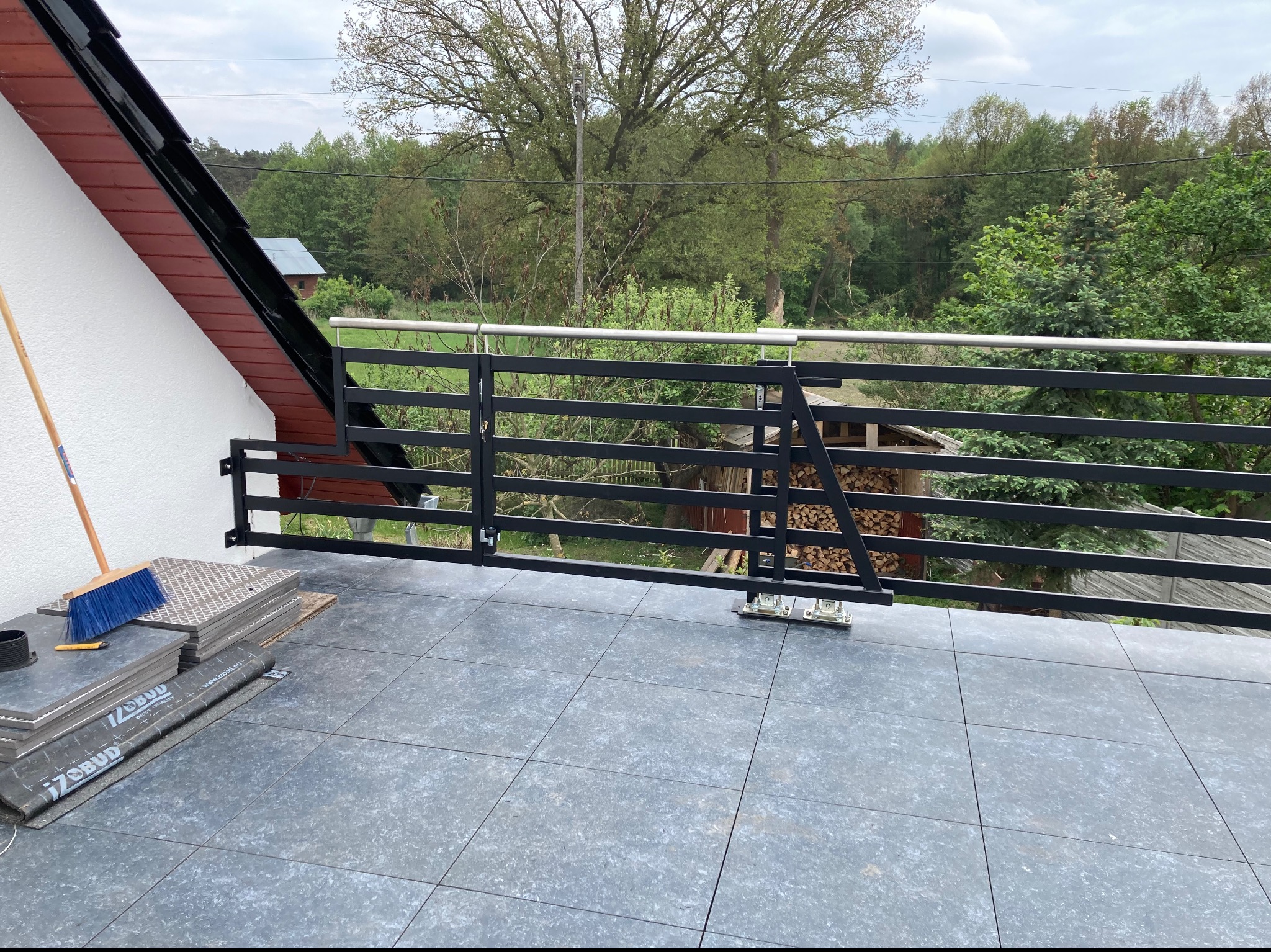 Częściowo zamontowana czarna metalowa balustrada z furtką na tarasie z szarych płytek, widoczne materiały budowlane i narzędzia, tło stanowi zielony krajobraz.