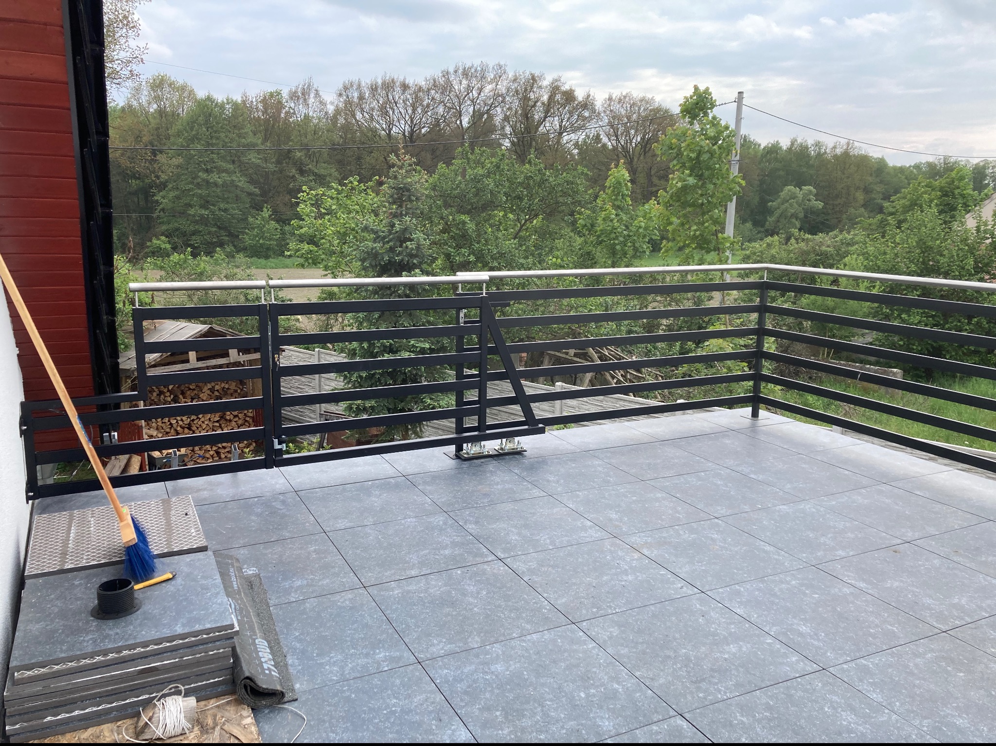 Nowo wykończony taras z szarymi płytkami, czarną metalową balustradą z elementami ze stali nierdzewnej oraz widokiem na zieleń, podczas prac wykończeniowych widoczna miotła i zapas płytek.