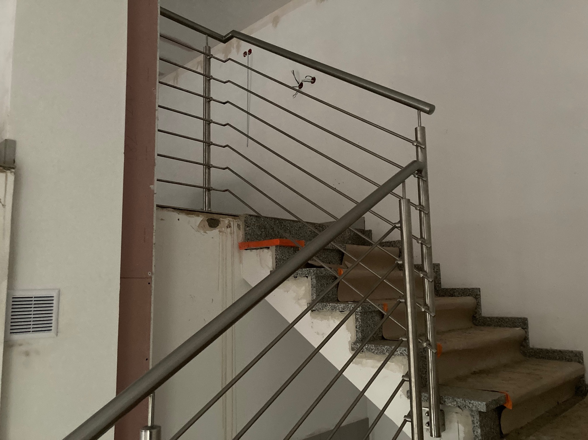 Wewnętrzne schody wykończone szarym kamieniem z zabezpieczonymi stopniami, z zamontowaną balustradą ze stali nierdzewnej z poziomymi prętami, widoczne niedokończone prace wykończeniowe ścian.