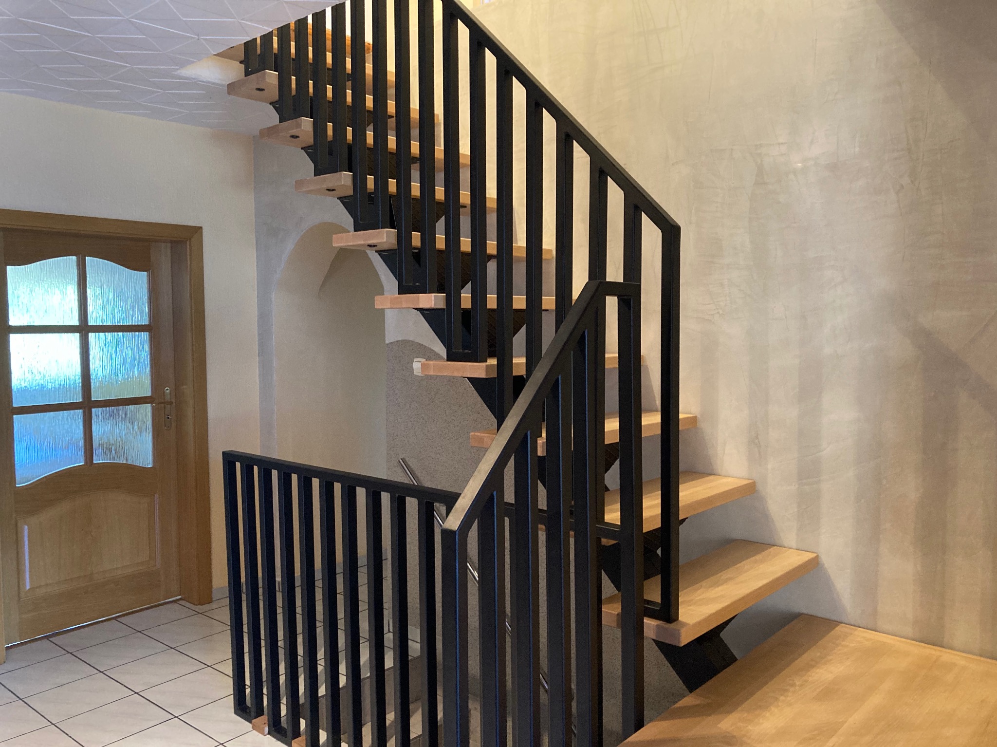 Nowoczesne schody z drewnianymi stopniami i czarną, minimalistyczną balustradą z pionowych prętów, widok z dołu na tle jasnej ściany i drzwi.